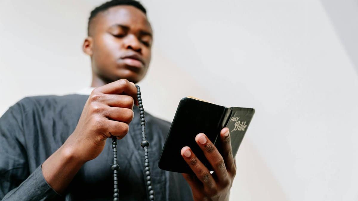 Focused young man holding a Bible and rosary beads in prayer at home.