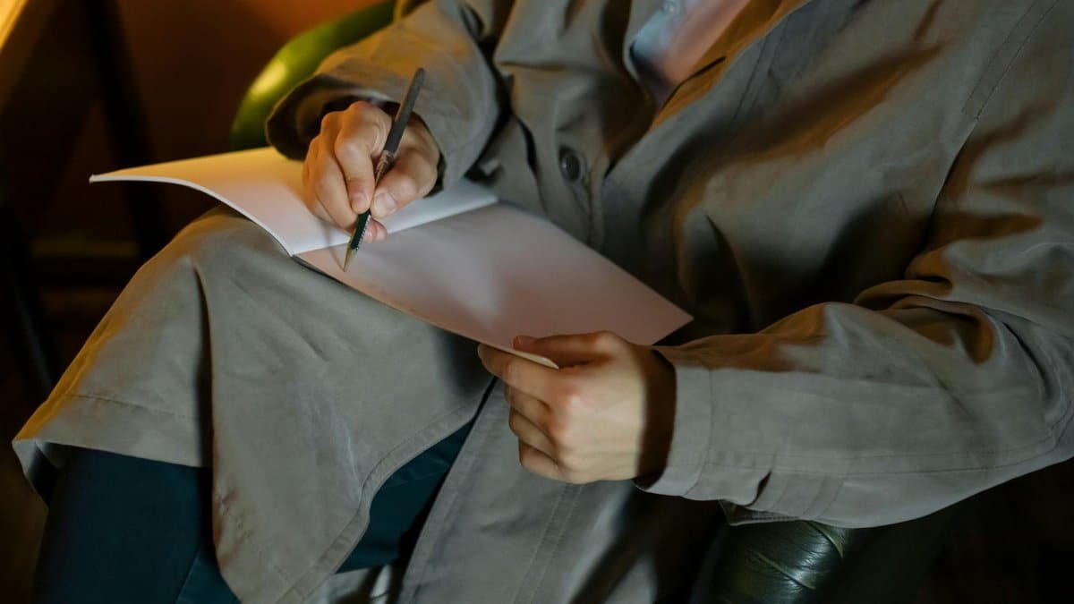 A person in a coat writes in a notebook in a warm, indoor environment, focused on creativity and thought.