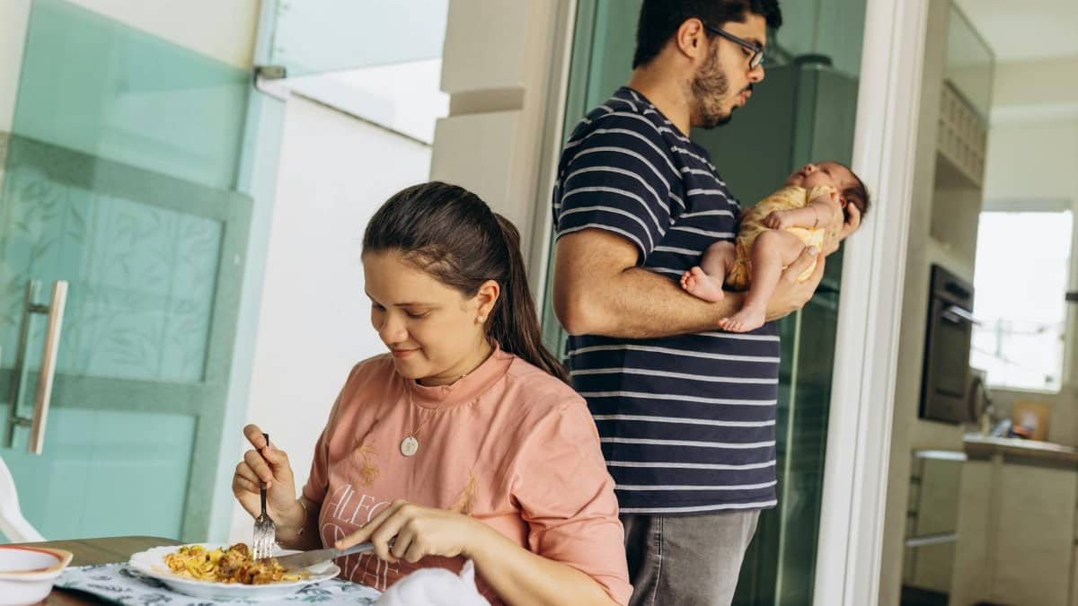 A woman enjoys a meal while a man cradles a baby, capturing family life.