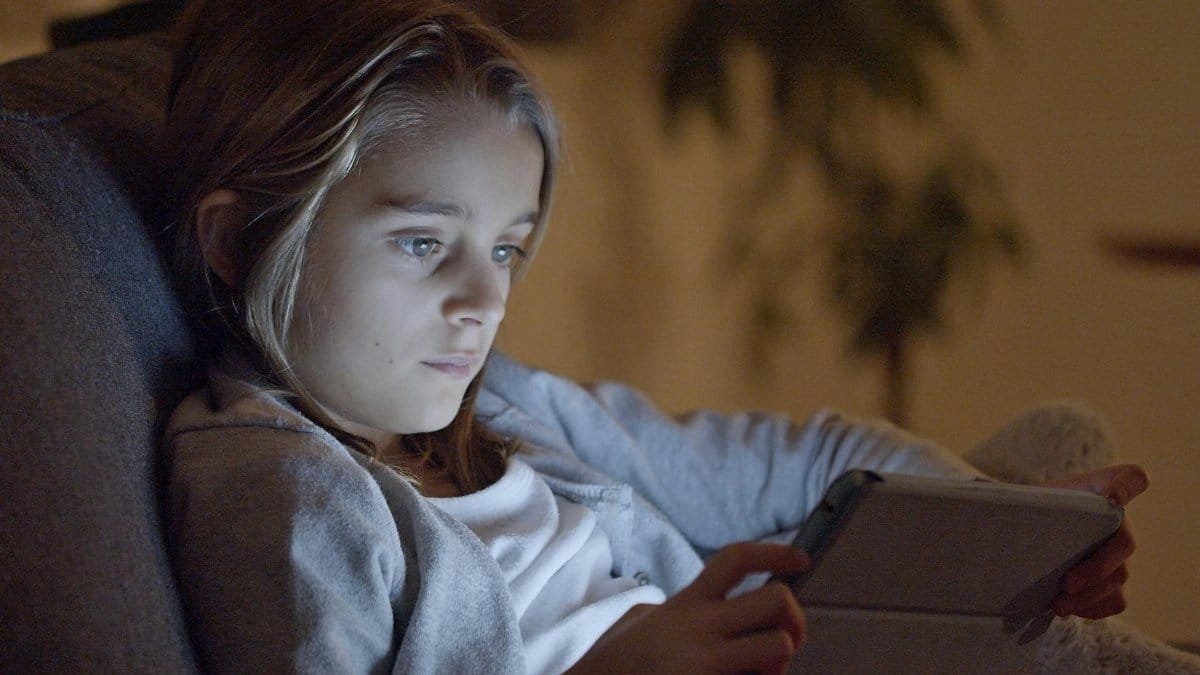 A young girl focuses on a digital tablet while relaxing on a couch at night.