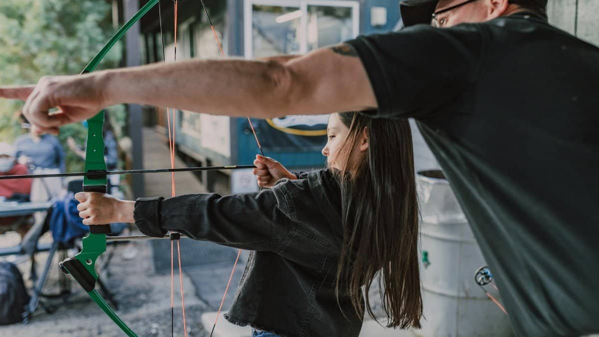 A young girl is learning archery with guidance from an adult man outdoors, focusing on target practice.