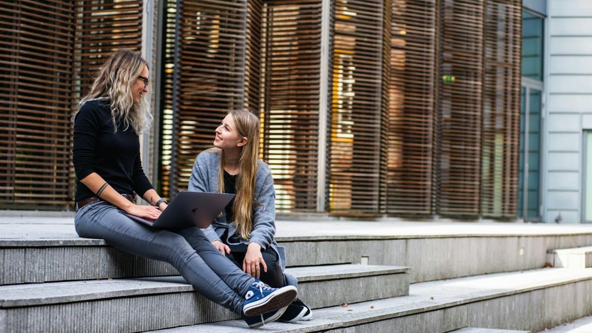 Two women sitting on steps with a laptop, discussing ideas in urban setting.
