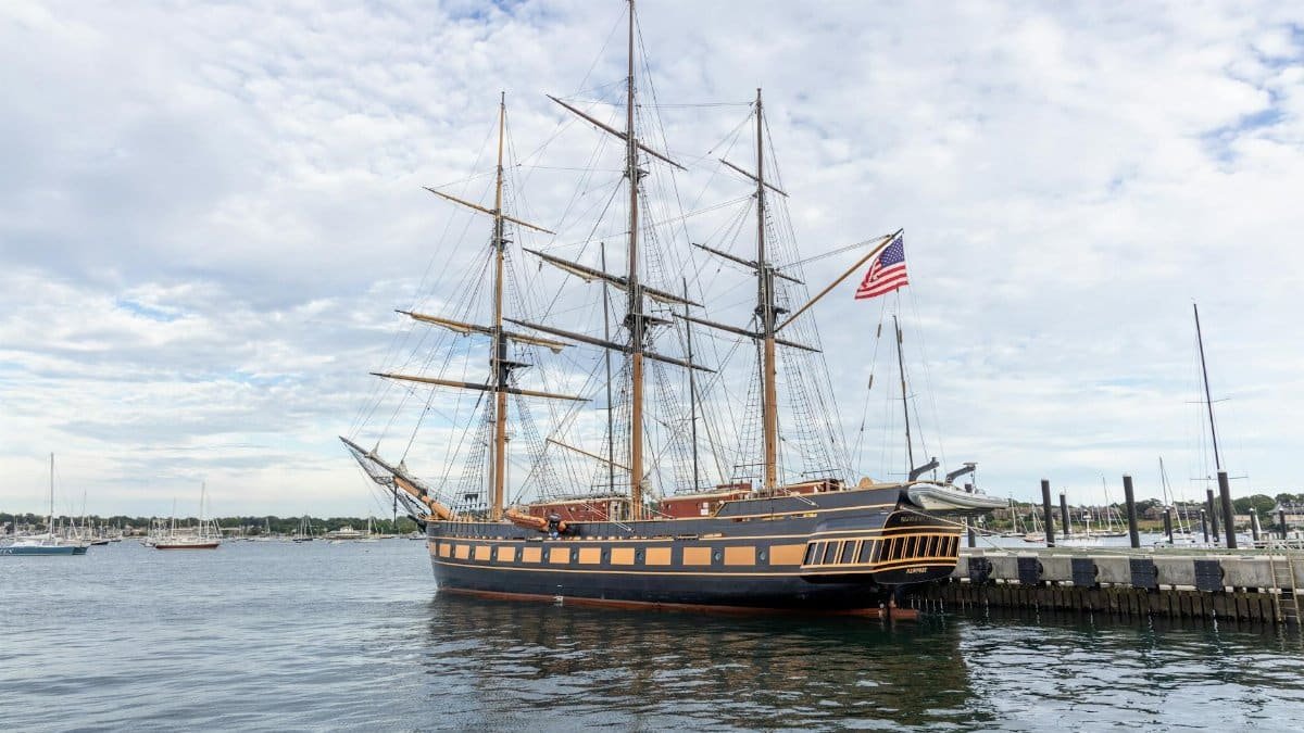 A majestic historic ship with an American flag docked in Newport, RI harbor, under a cloudy sky.