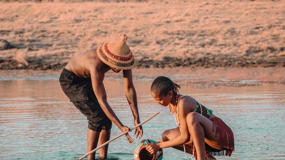 Two people in traditional attire gathering water from a river in a rural setting.