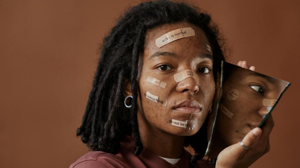 A woman holds a broken mirror with positive messages on her face, reflecting self-acceptance.