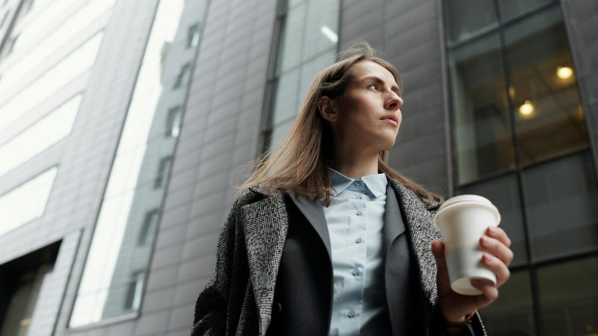 Professional woman in a city street holding a coffee cup, gazing upwards.