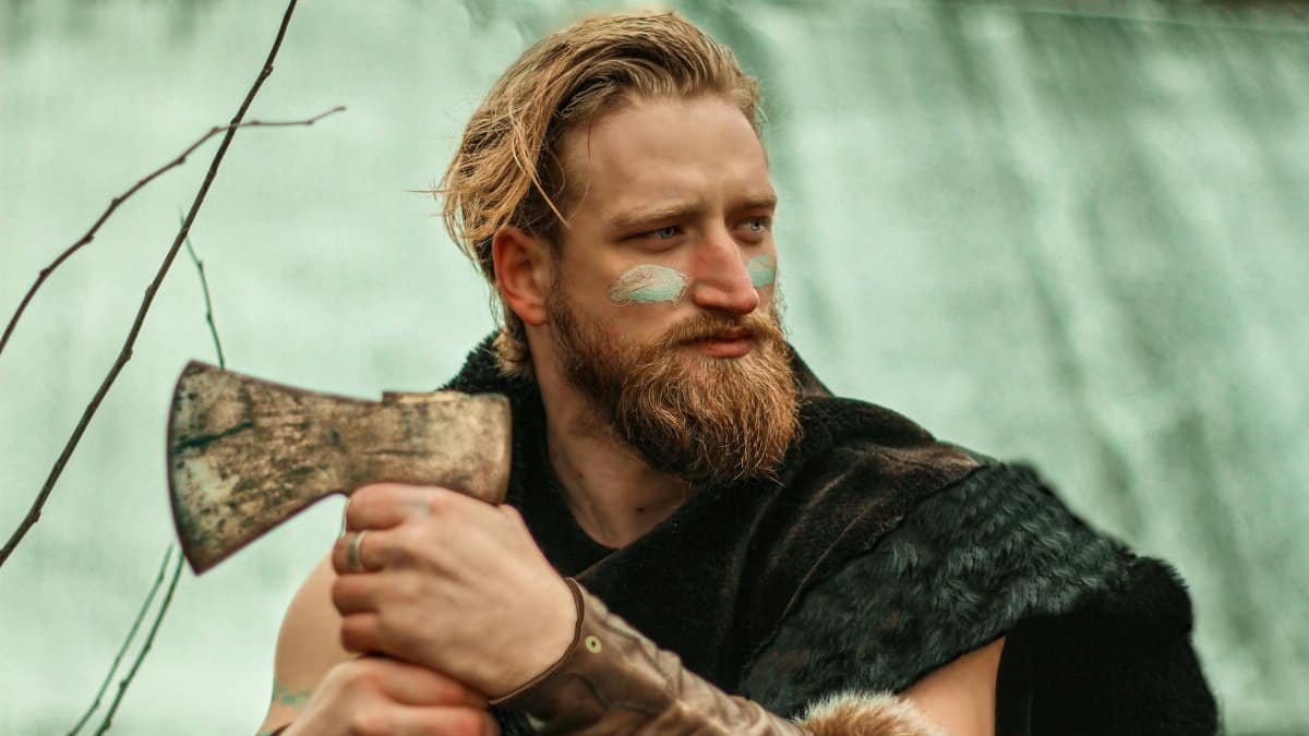 Bearded man dressed as a Viking with face paint, holding an axe outdoors.