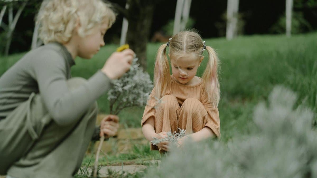 Two children engage in outdoor play and exploration, interacting with plants in a grassy garden.