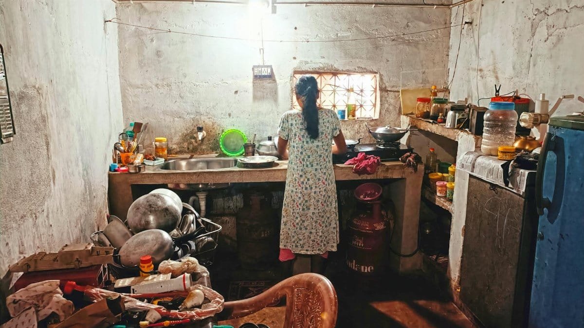 A woman in a floral dress cooks in a rustic Indian kitchen, offering a glimpse of daily life.