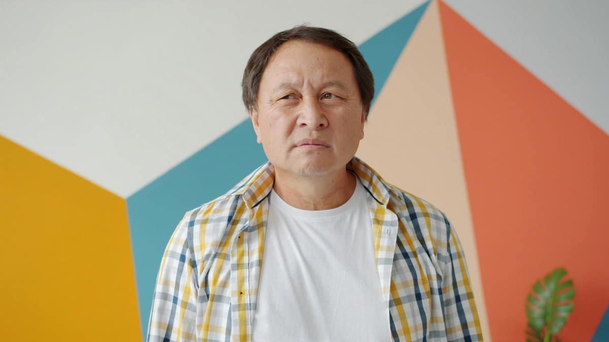 Middle-aged man in checkered shirt standing indoors with colorful geometric wall background.