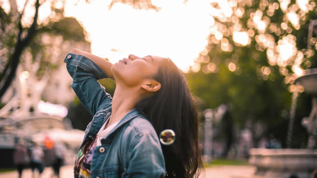 Low angle of happy brunette in denim jacket standing alone with eyes closed and head threw back enjoying life and youth among soap bubbles against blurred urban environment in sunny day