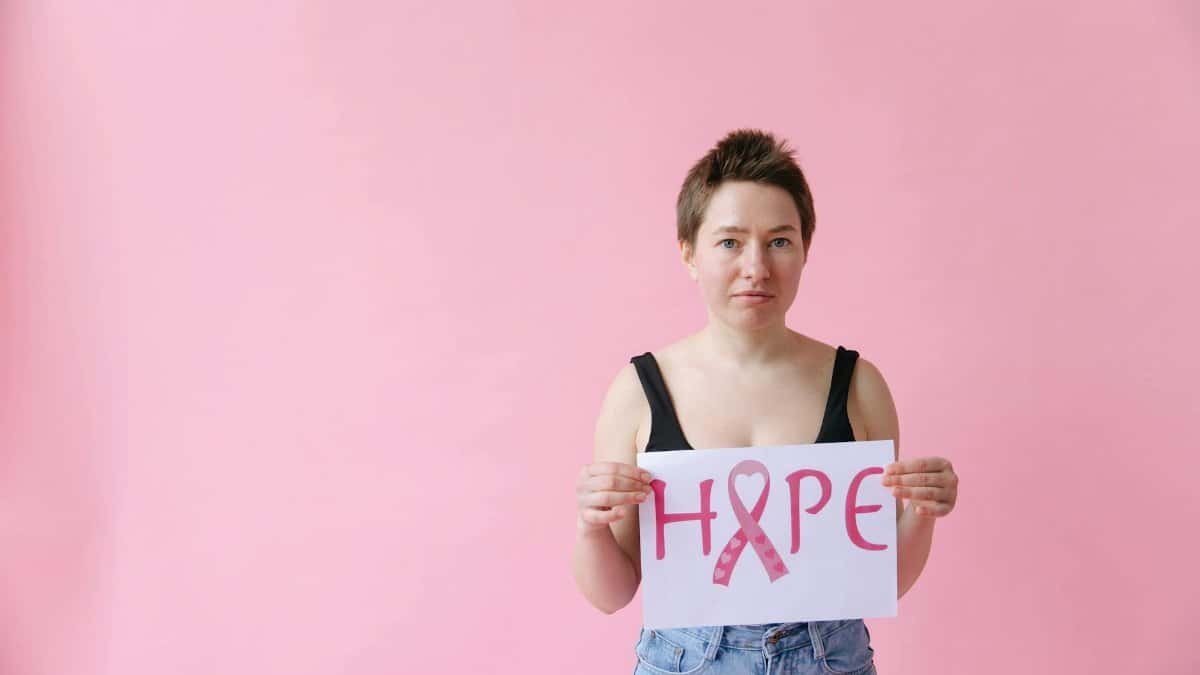 Woman holding a pink ribbon hope sign on pink background, symbolizing breast cancer awareness.