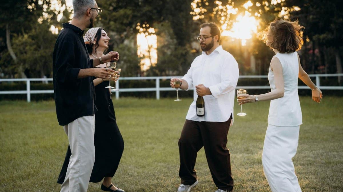 Four adults enjoying an outdoor celebration, dancing and holding drinks as the sun sets.