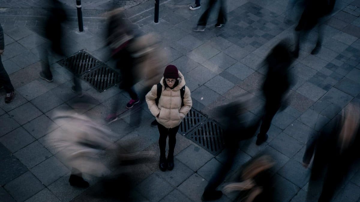 A woman standing still on a city sidewalk as blurred people walk around her, symbolizing isolation amidst urban hustle.