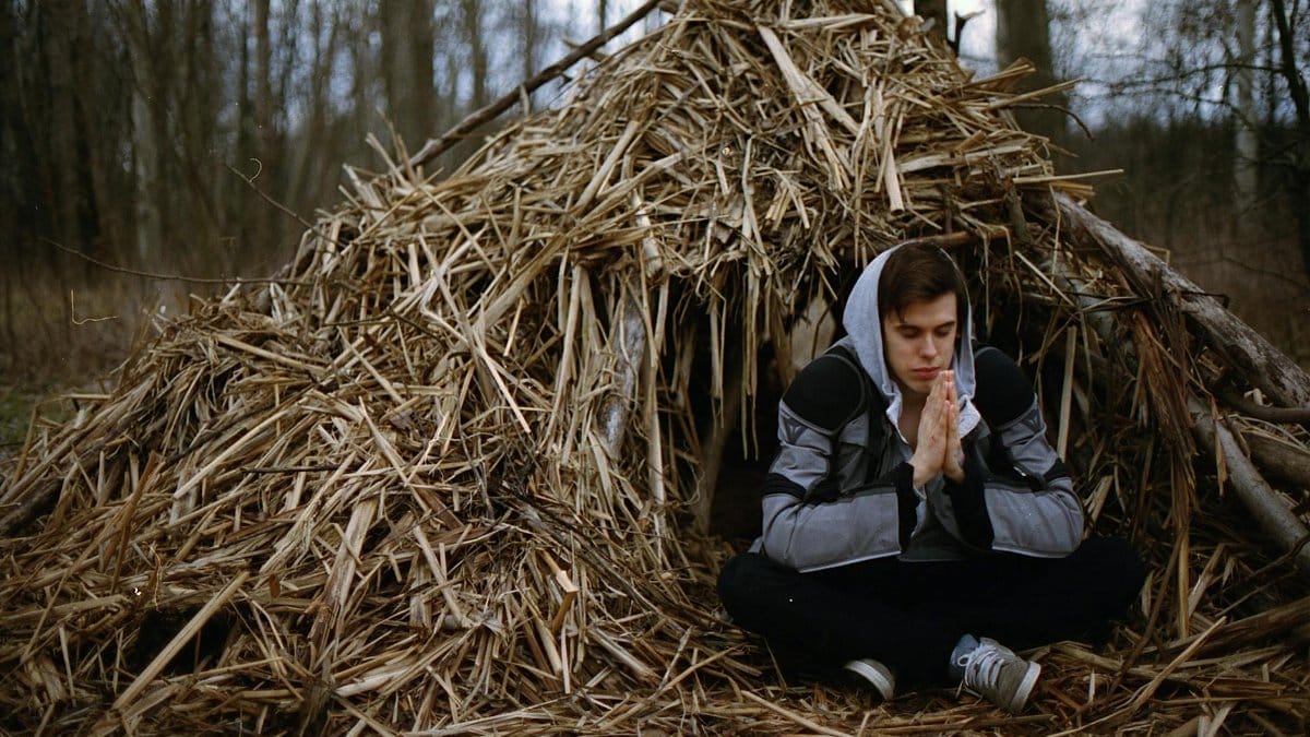 Adult male meditating outdoors by a rustic hay hut in a serene forest setting.