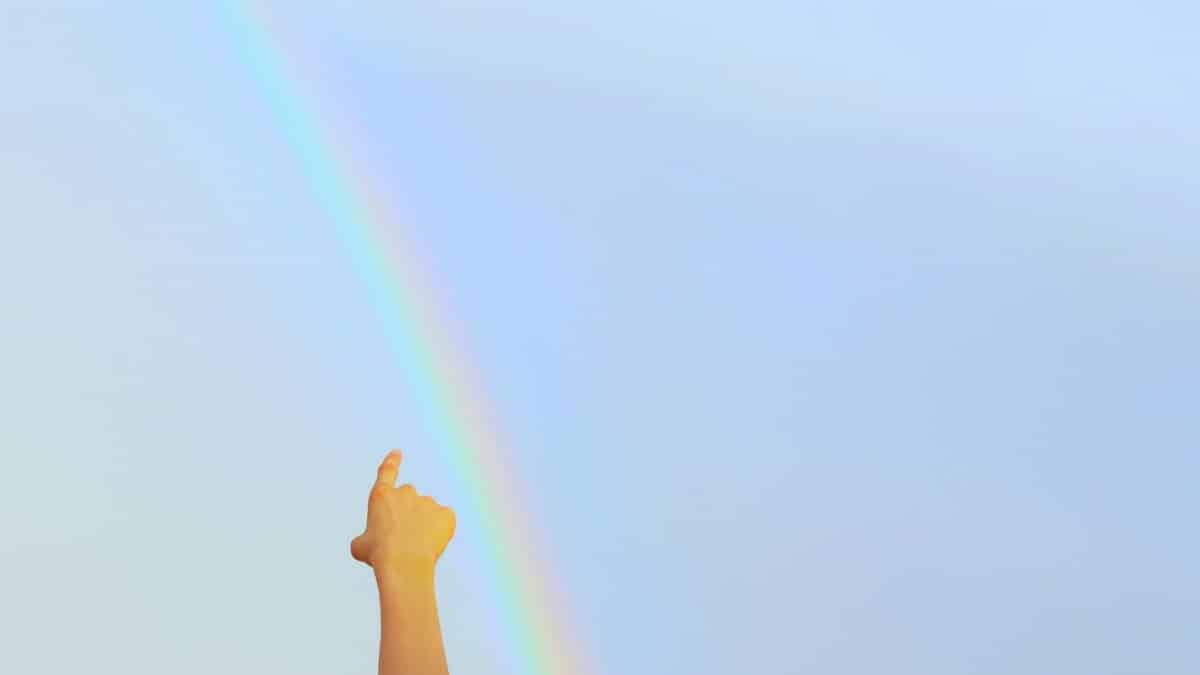 A child's hand pointing towards a bright rainbow against a clear blue sky, symbolizing hope and wonder.