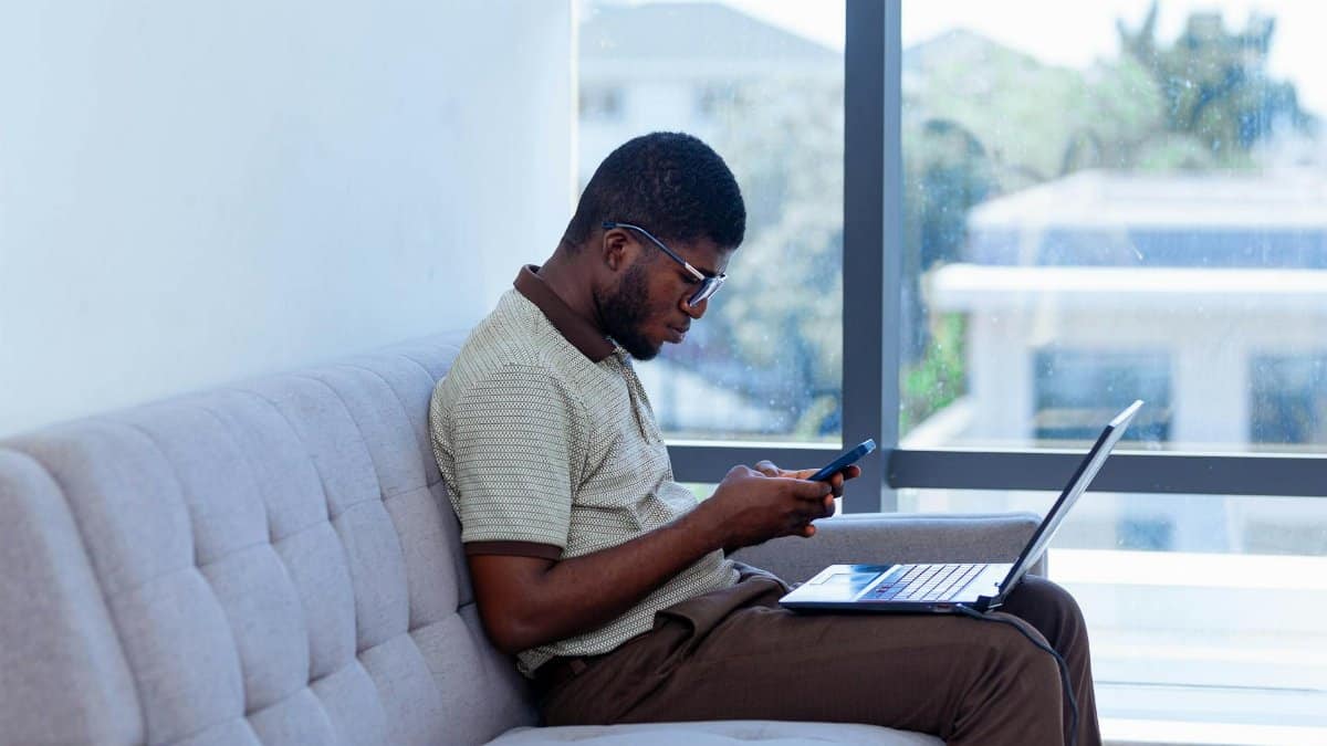 Man multitasking with laptop and smartphone indoors, showcasing remote work lifestyle.