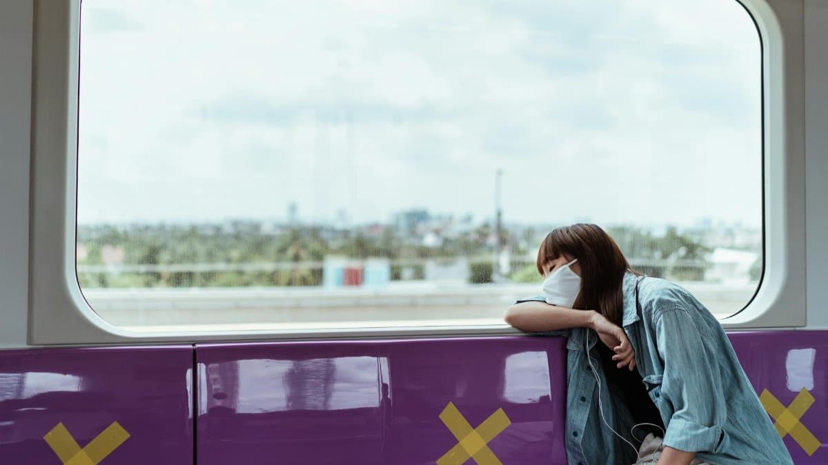 A woman wearing a face mask rests on a train seat, observing social distancing measures.