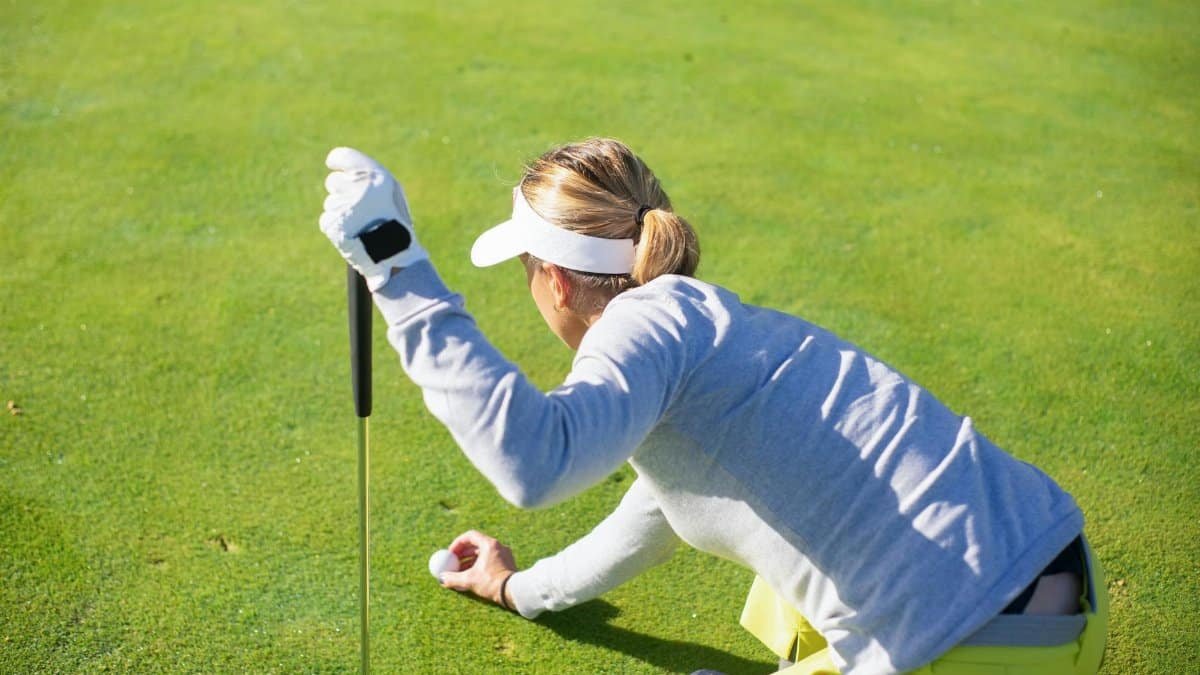 Female golfer in a gray sweater prepares to putt on a bright day outdoors.