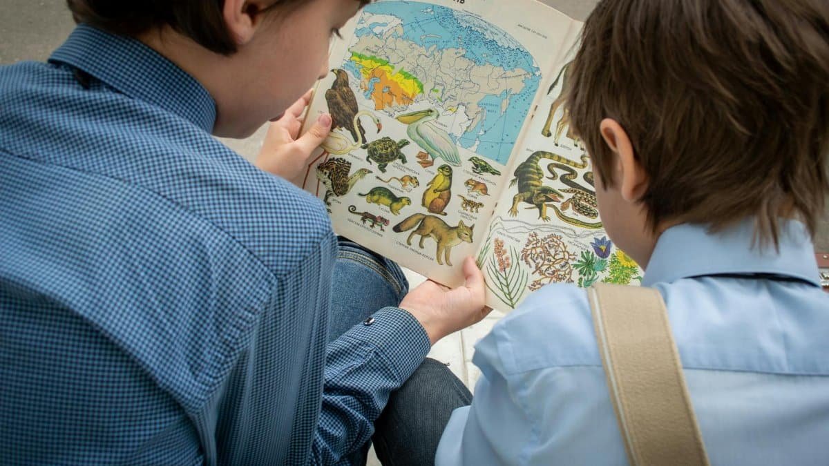 Two children outdoors reading an illustrated nature book, focused on learning animals and geography.