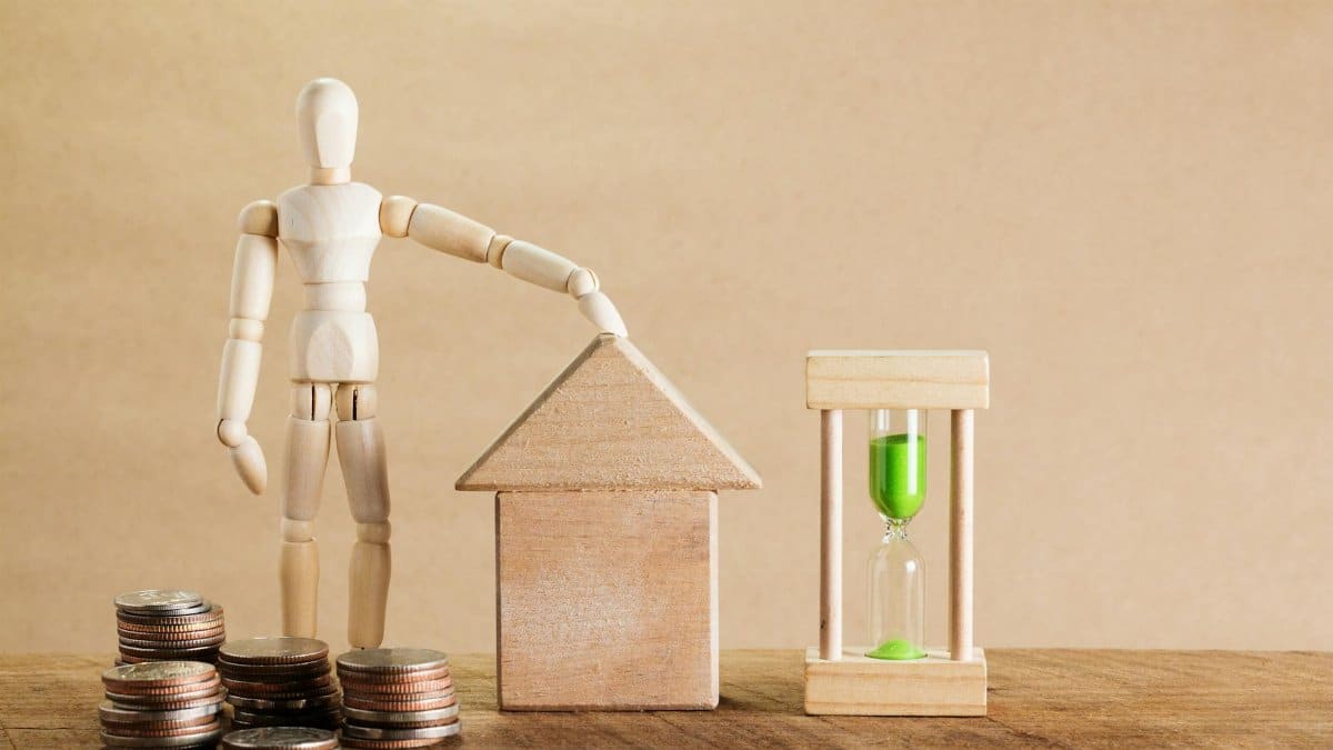 Wooden mannequin with house model, coins, and hourglass symbolizing financial planning.