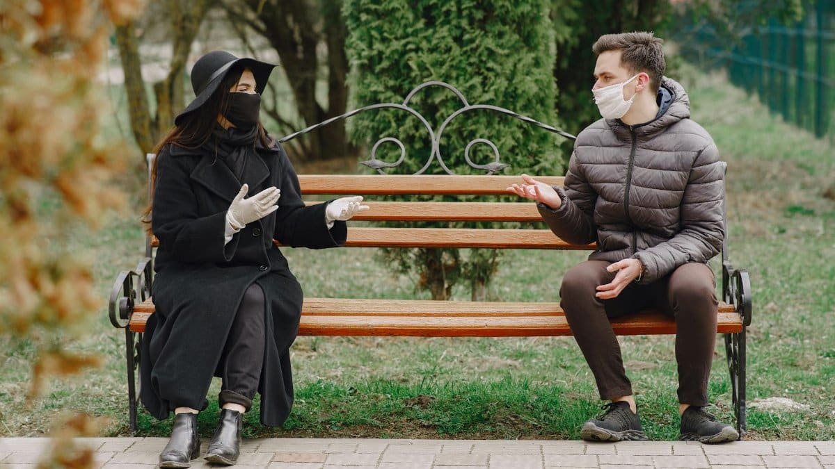 Serious unrecognizable couple in warm casual clothes wearing protective latex gloves and medical masks sitting on bench in spring park and keeping social distance