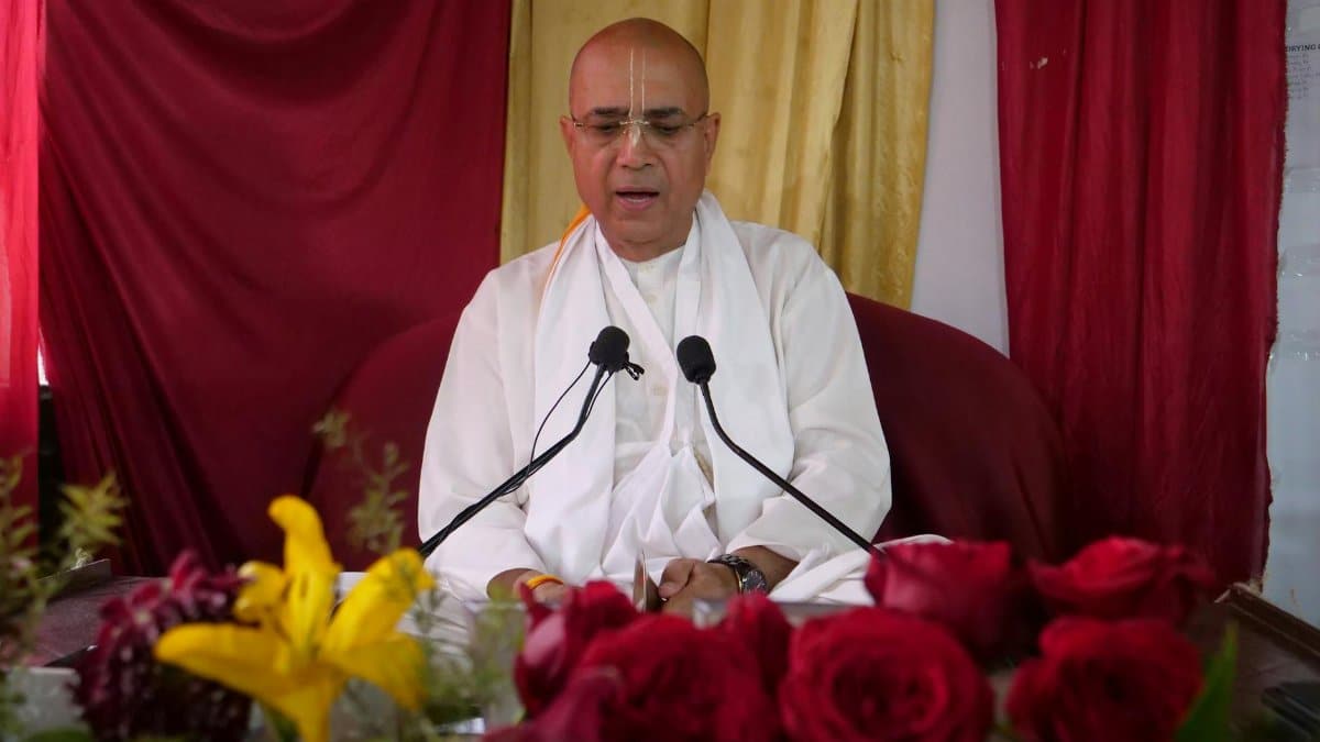 A senior spiritual leader delivers a religious sermon surrounded by colorful flowers and microphones.