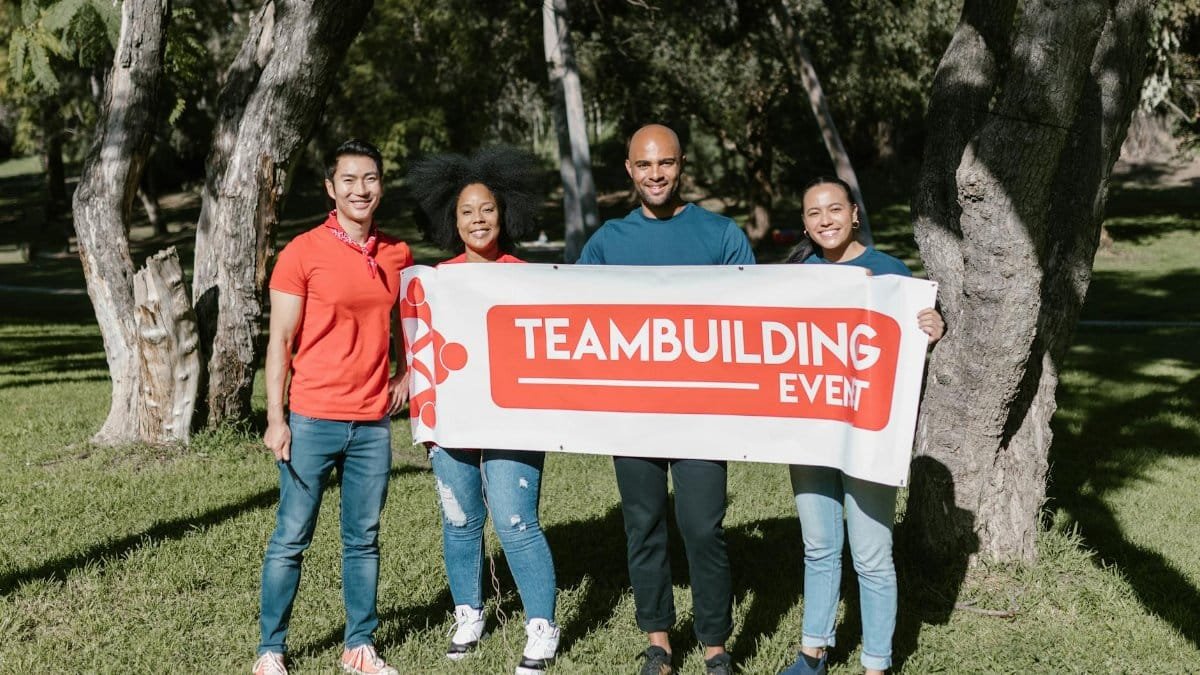 Group of four diverse adults holding a team building banner in a sunny park.