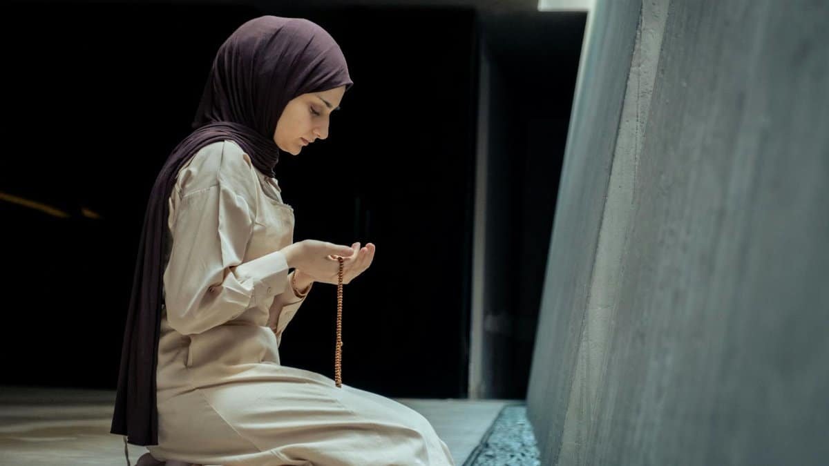 A woman in hijab kneels in prayer with beads inside a peaceful mosque environment.