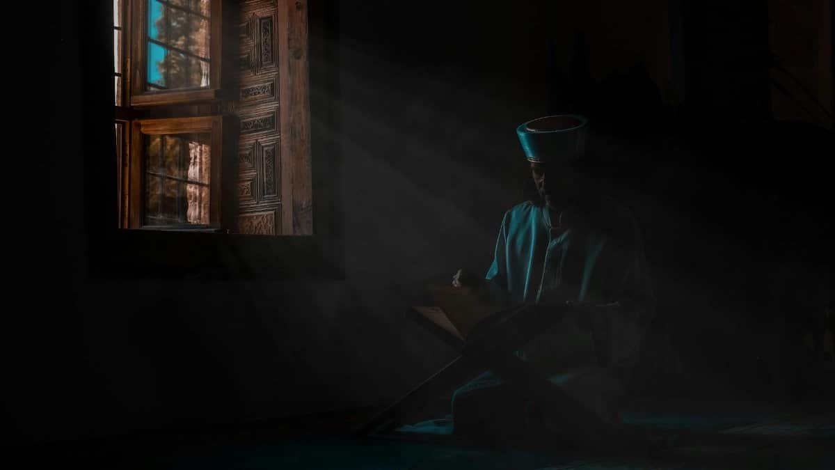 Man in traditional clothing reading by the window, enveloped in spiritual ambiance.