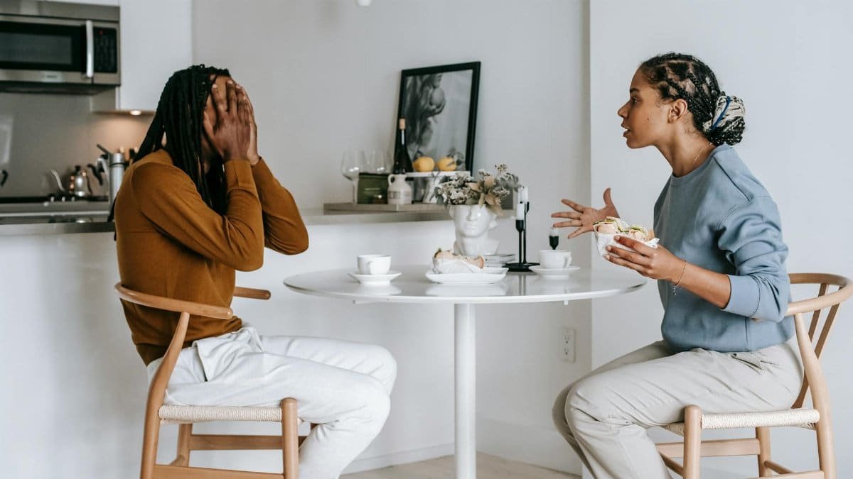 Side view of expressive Hispanic female in casual clothes arguing with African American boyfriend covering face with hands while sitting at table in kitchen at home
