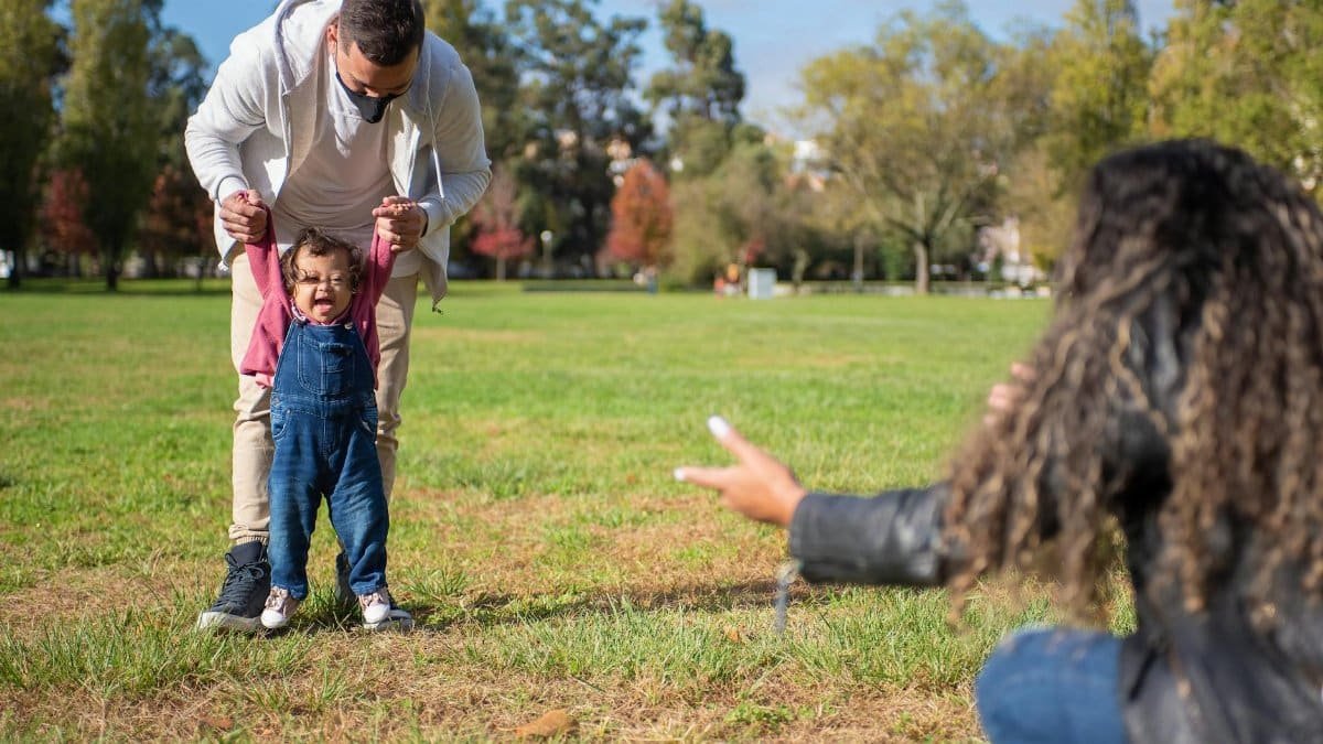Father supporting his baby taking first steps while mother reaches out in a sunny park.