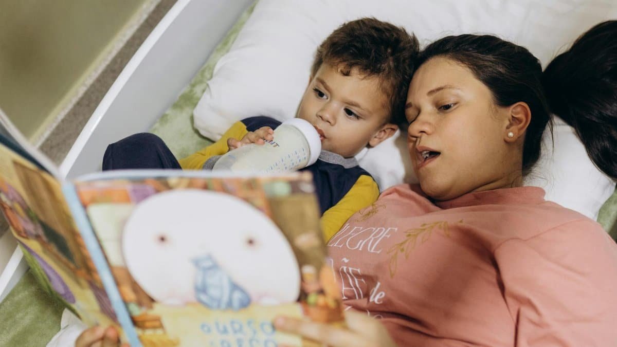 A mother reads a bedtime story to her child while he drinks milk, creating a cozy and bonding moment.