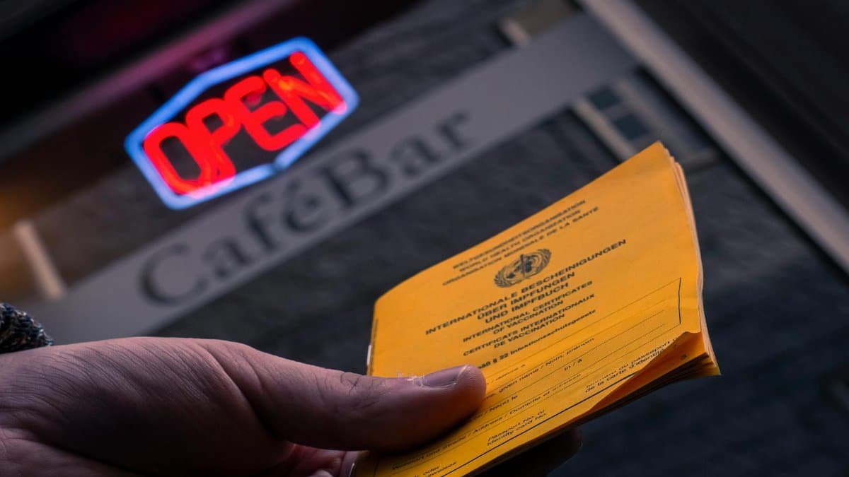 Close-up of a hand holding a vaccination card in front of a cafe bar with an open sign.