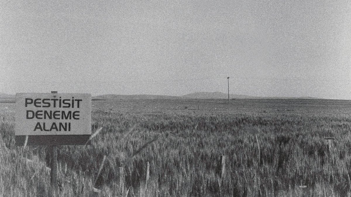 A black and white photo of a rural field with a sign that reads 'PESTISIT DENEME ALANI.'