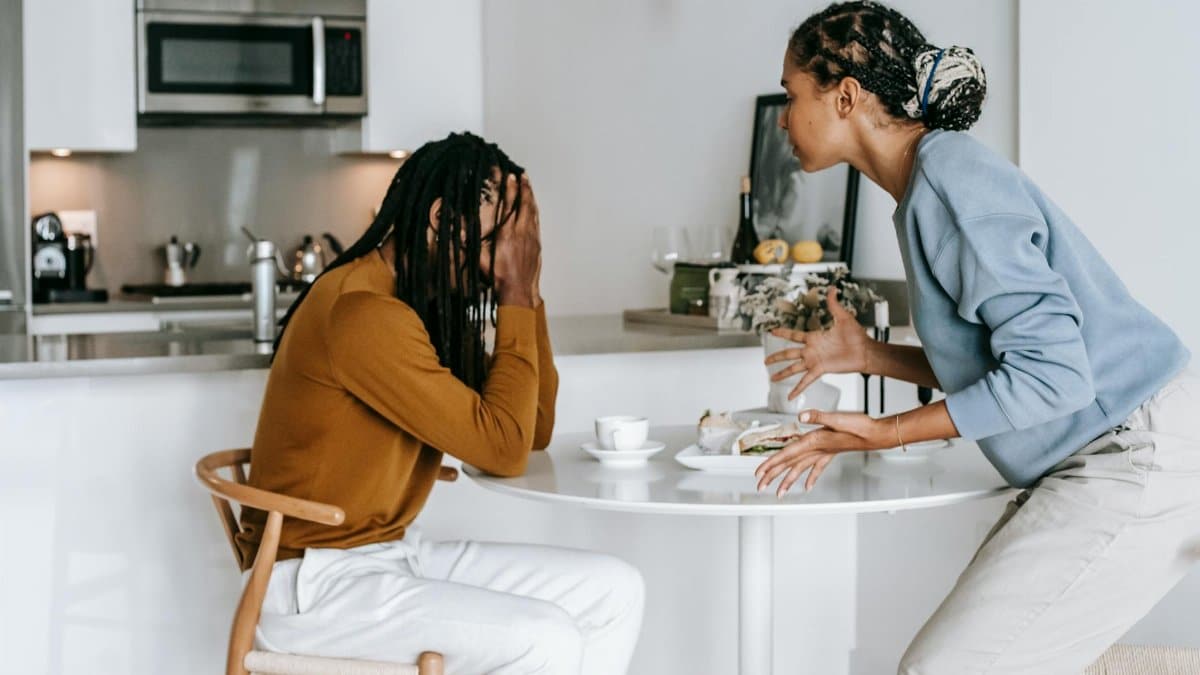 Young black man sitting at table with hands on head and having conflict with standing near table woman in light kitchen