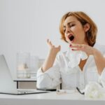 A woman in white yawning while working at a desk, indicating fatigue.