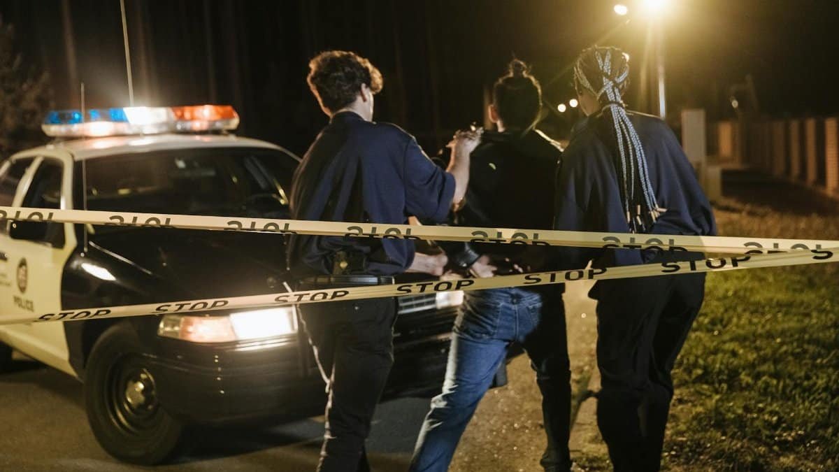 Nighttime arrest scene with police officers and patrol car behind caution tape.