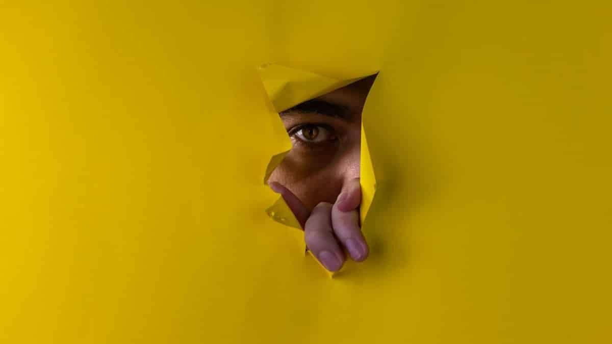 A close-up of an eye peeking through torn yellow paper, creating a feeling of intrigue and curiosity.
