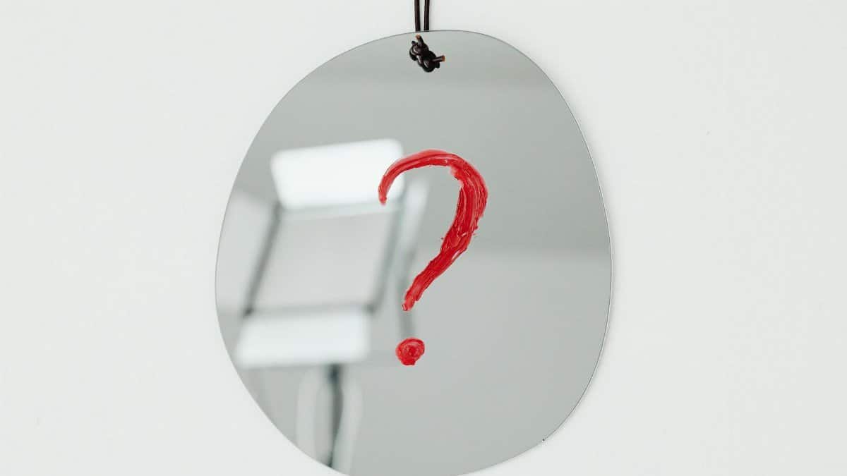 An oval mirror with a red question mark painted on it, hanging against a neutral background.