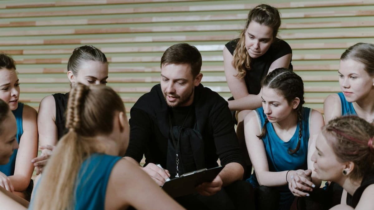 A coach discusses strategy with a women's team seated in a gym setting.