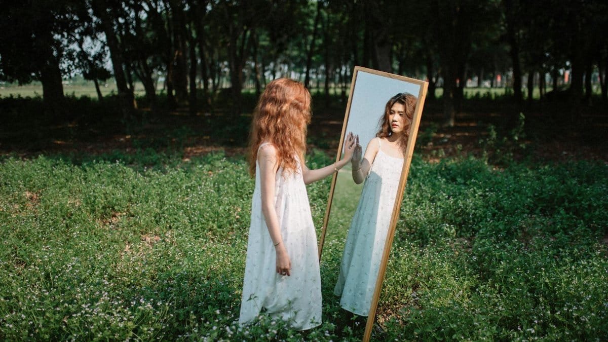 Young woman in a white dress gazing at her reflection in a mirror amid a lush green park setting.