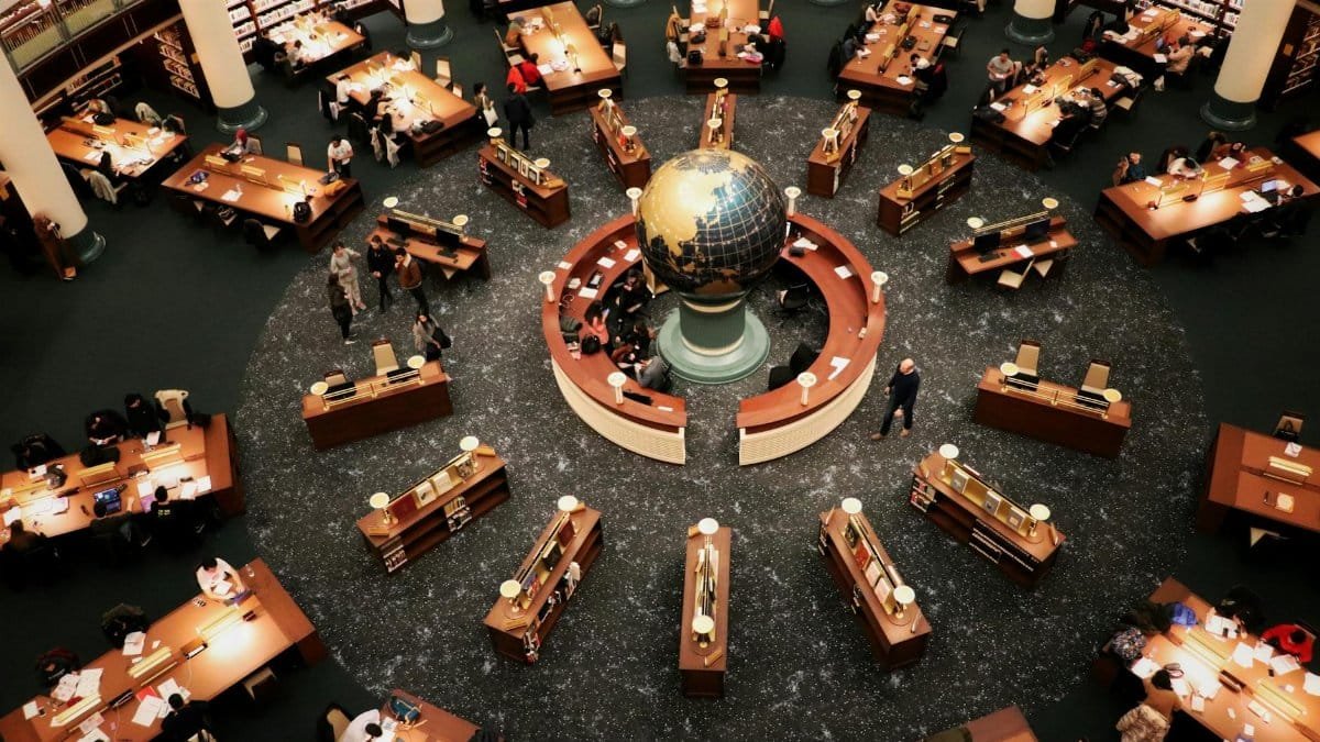 Busy library interior with a large globe centerpiece and people working at wooden tables. High angle view.
