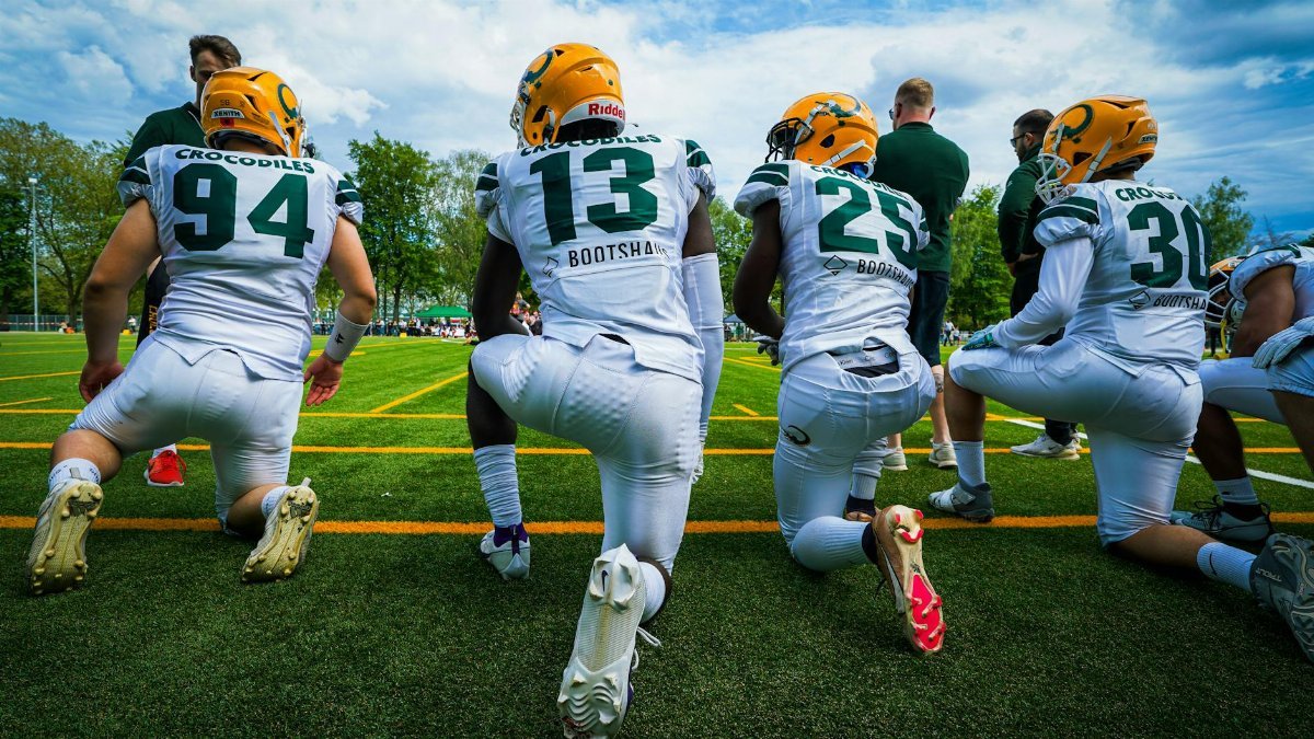 A group of American football players wearing helmets and sports gear kneeling on a green field.