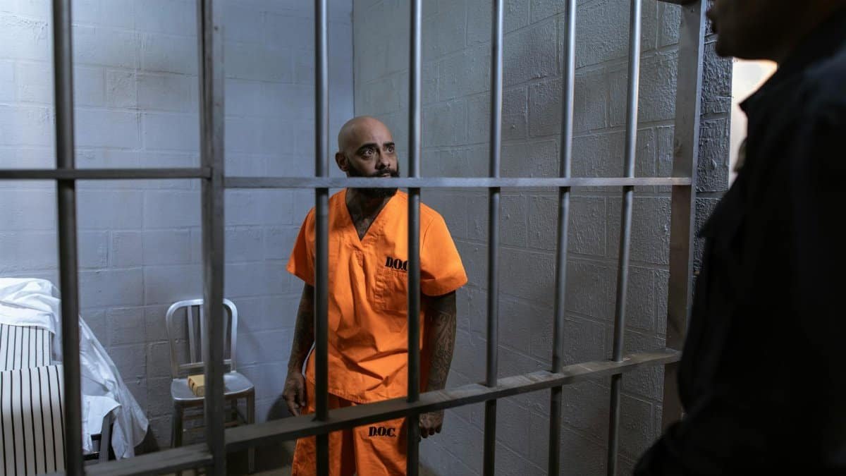 An inmate in an orange jumpsuit stands behind bars with a guard outside the cell.