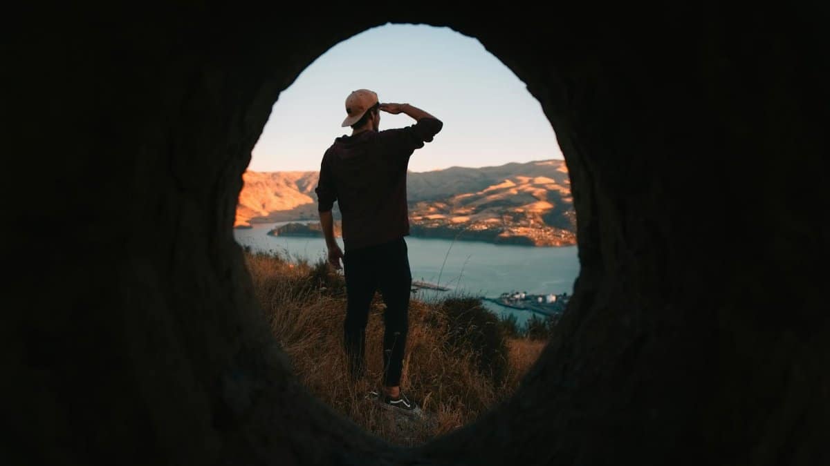 Man in a baseball cap observes a scenic coastal landscape through a tunnel at sunset.