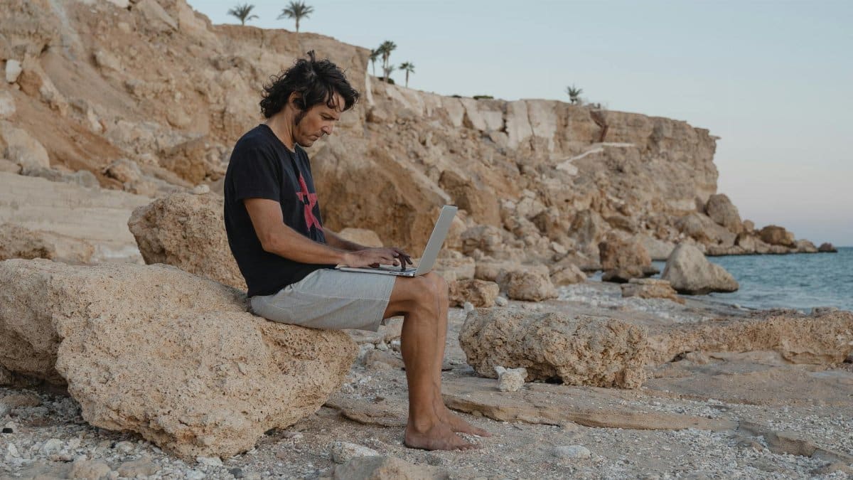 Man sits on rocks with laptop by the beach, enjoying remote work under a serene sunset.