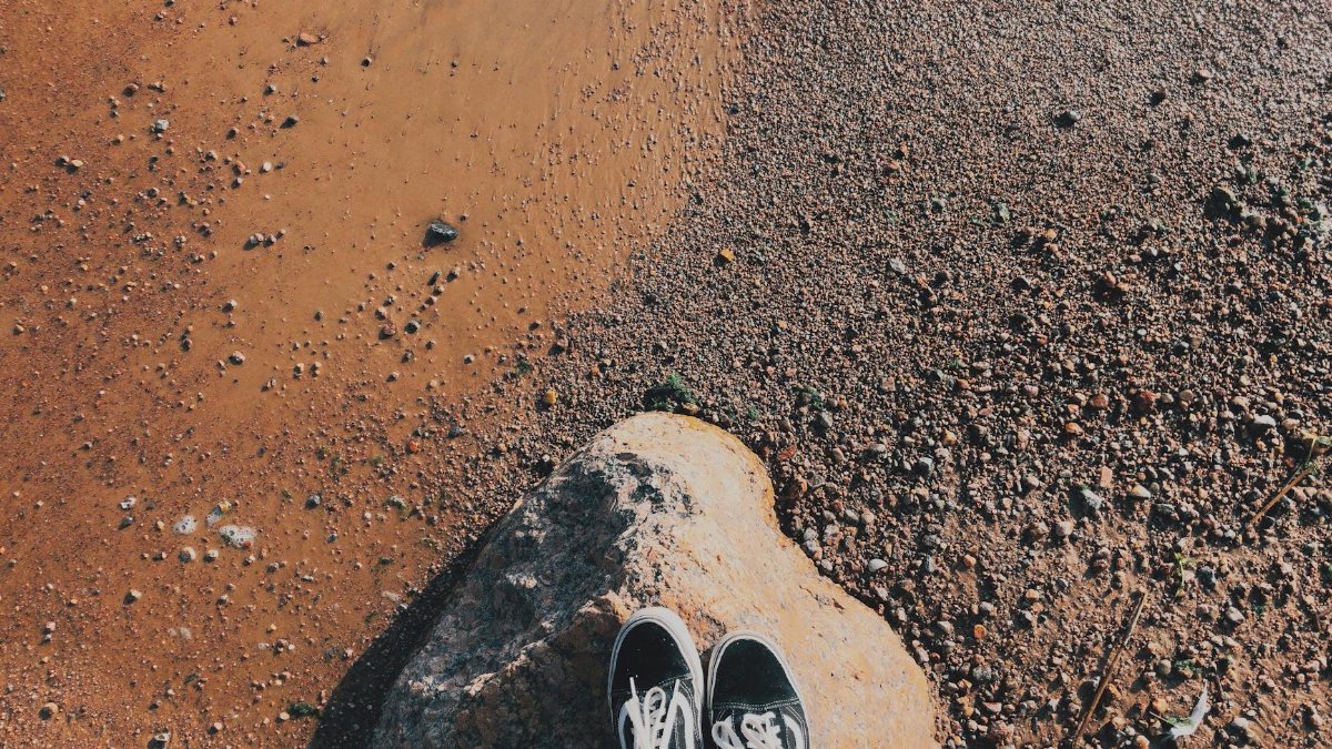 Person standing on rock by sandy beach with shoes. Ideal for travel and lifestyle themes.