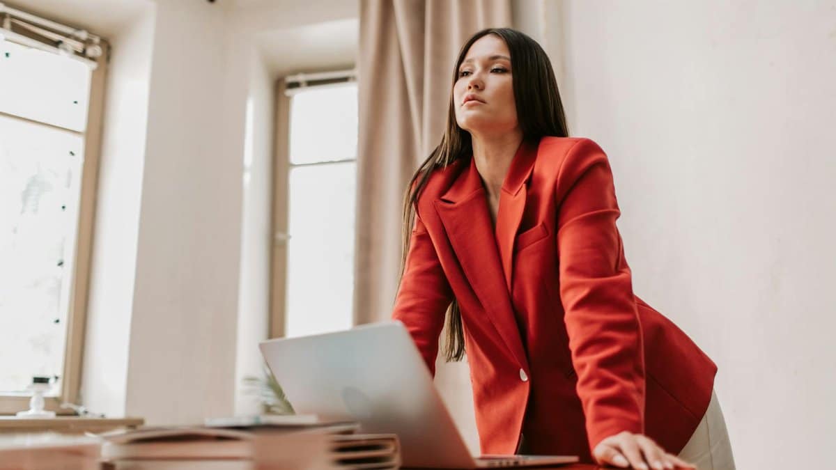 Businesswoman in a red blazer leaning over a desk with a laptop, exuding confidence.