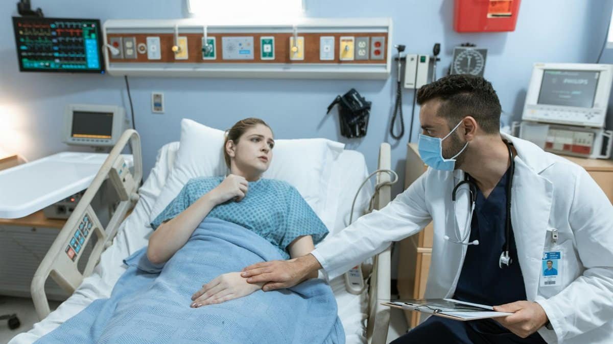 Male doctor with face mask comforts female patient in hospital bed with medical equipment.